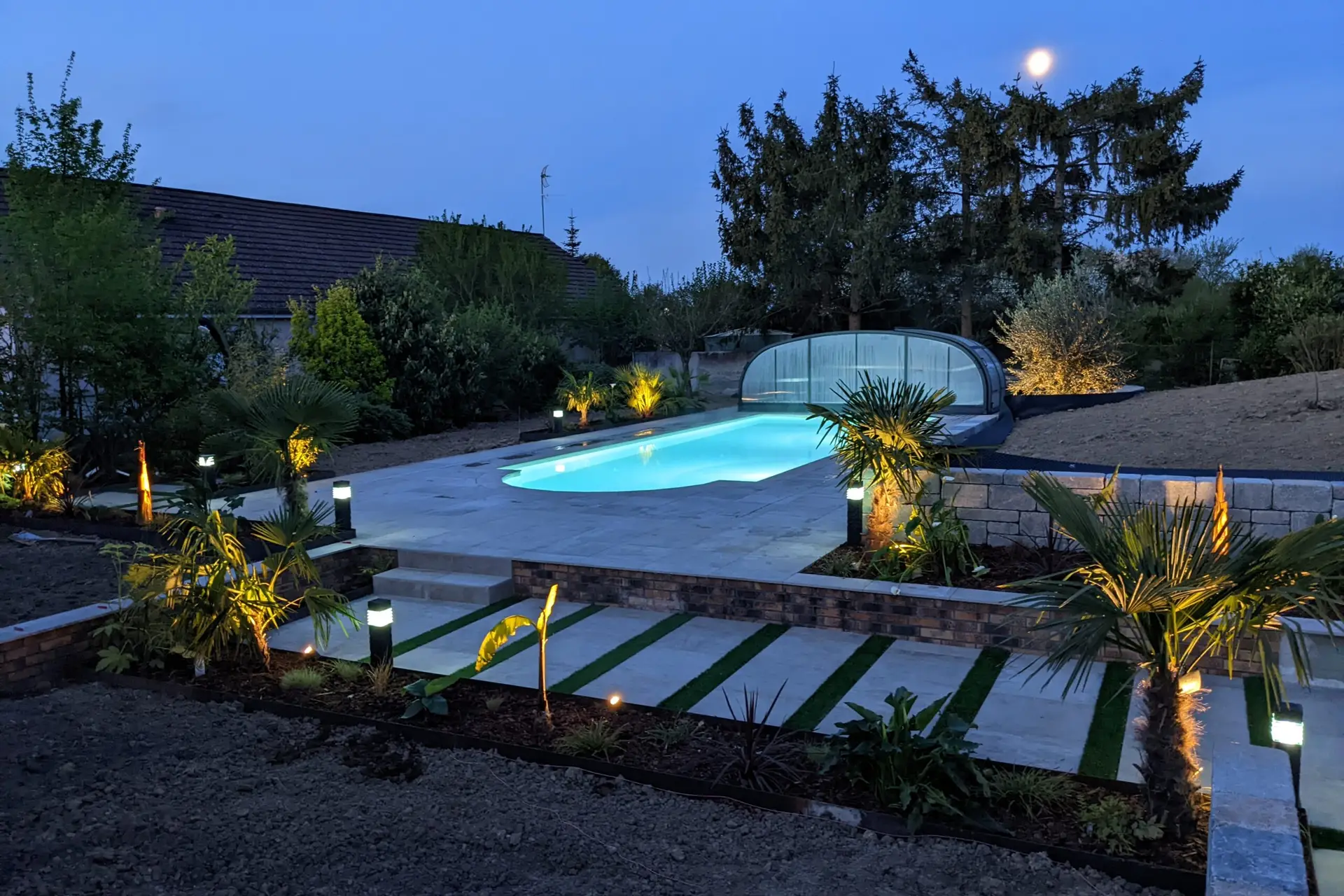 Jardin realise par EV2M a la tombee de la nuit : piscine eclairee, terrasse en dalles, muret en pierre, palmiers et eclairage paysager